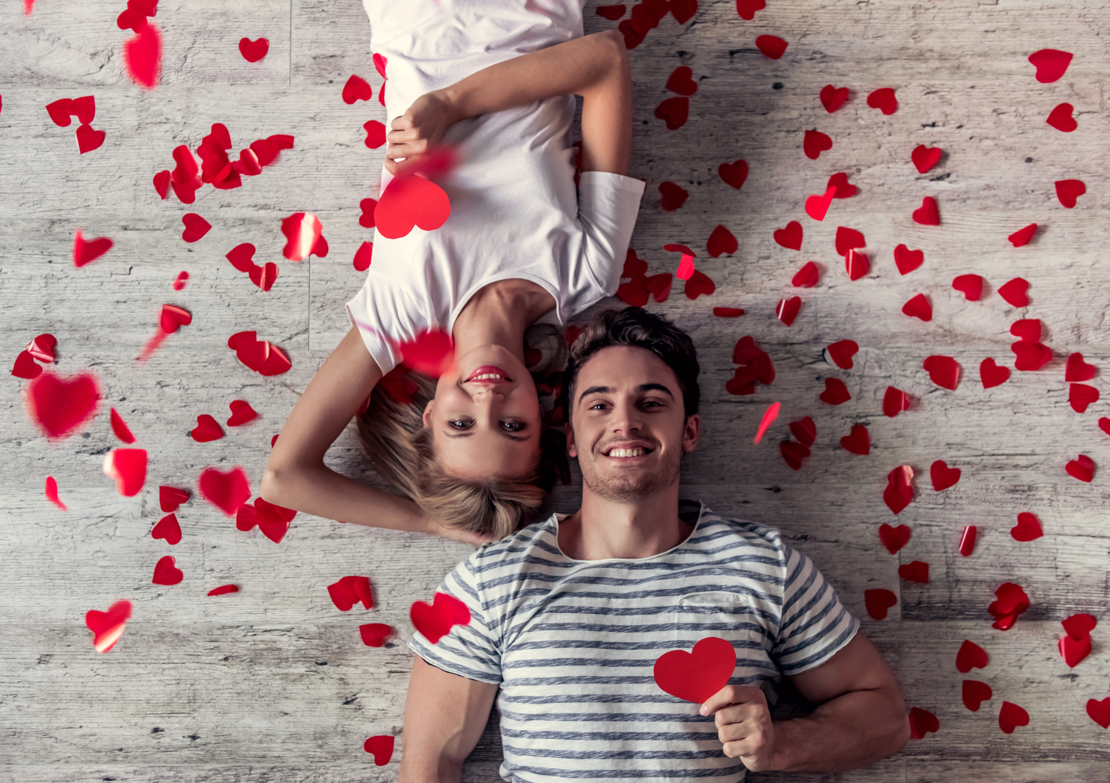 Romantic young couple Top view of beautiful young couple holding red paper hearts, looking at camera and smiling while lying on the floor