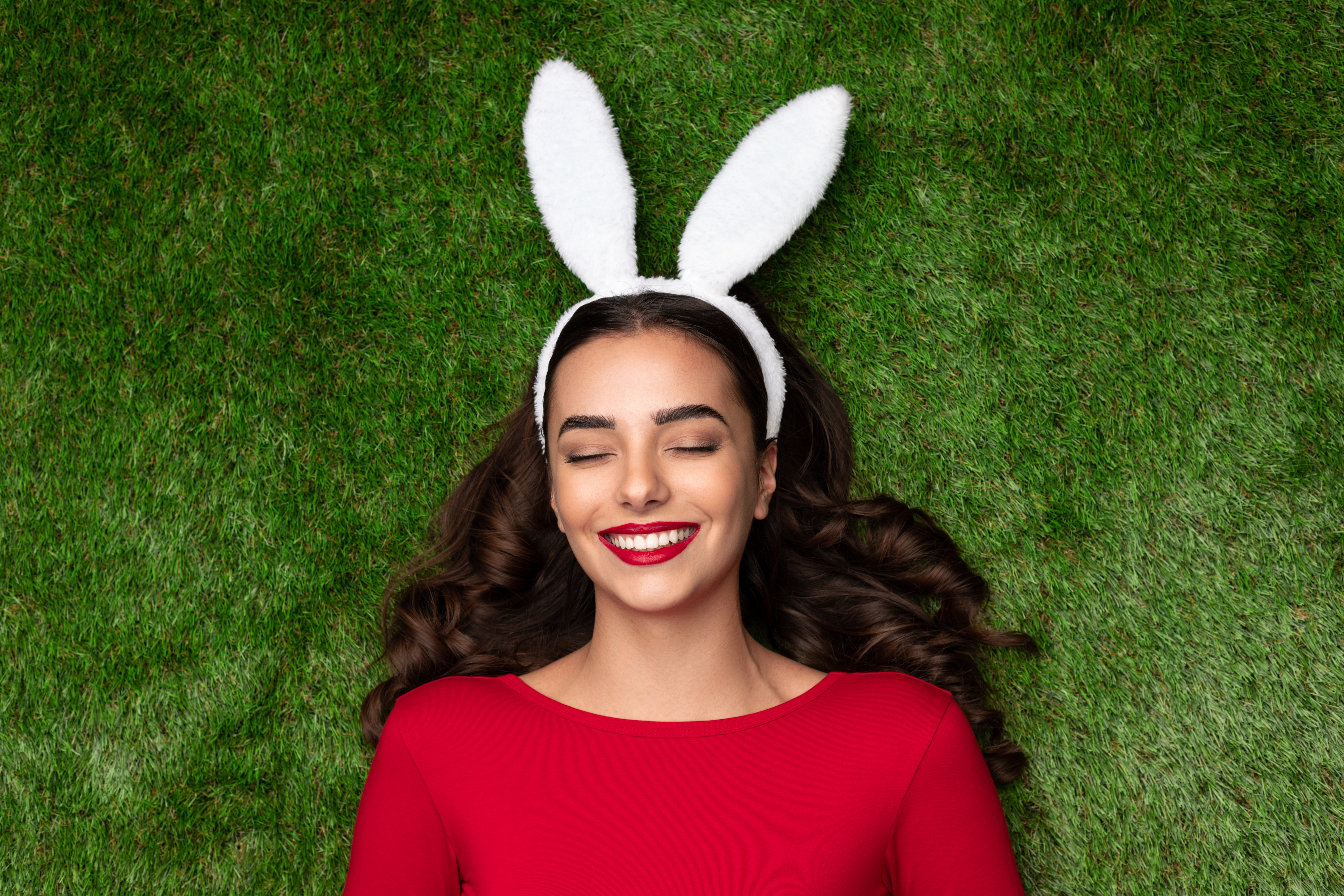 Adorable brunette in red dress and white bunny ears lying with eyes closed on green lawn