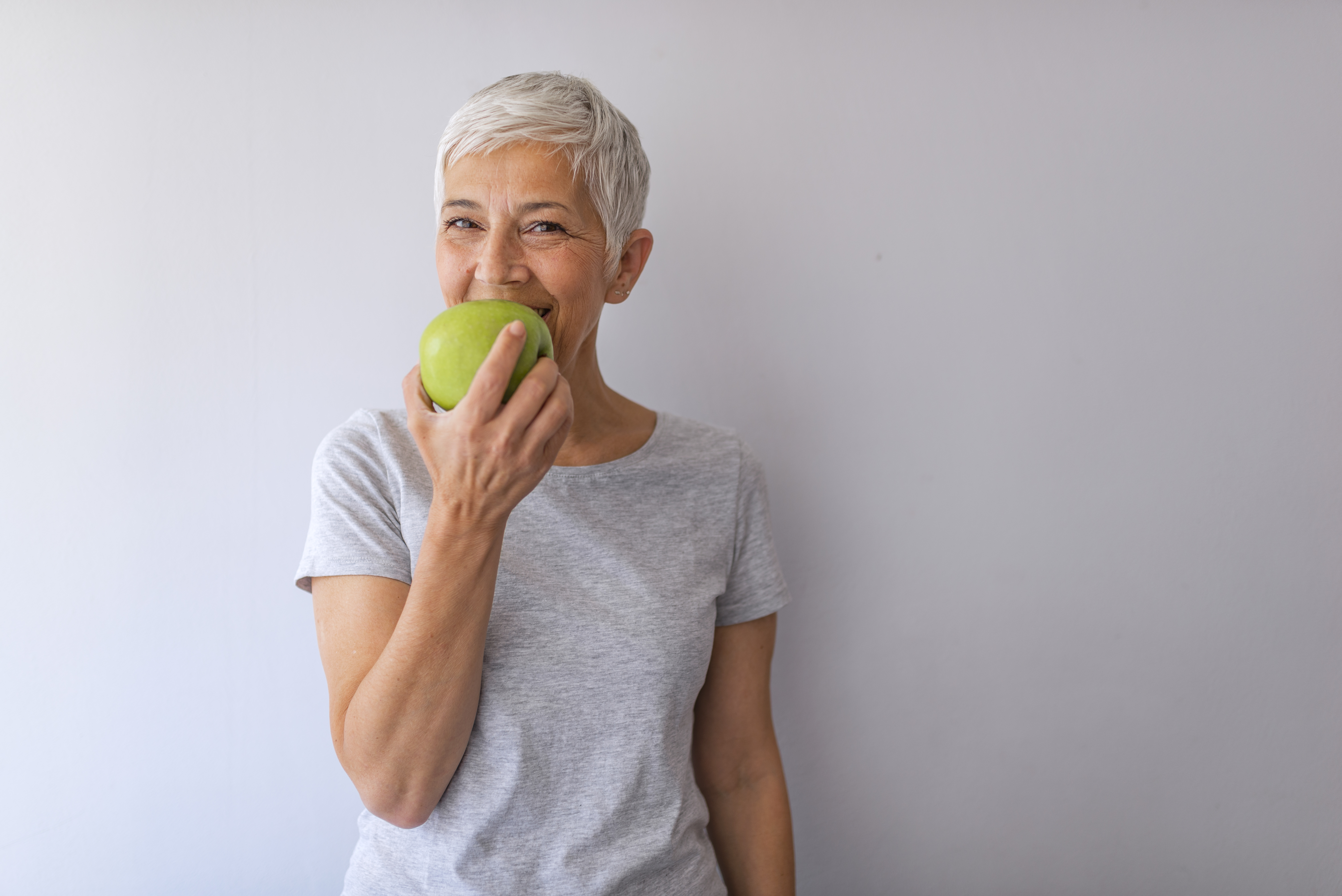 Beautiful senior woman over grey wall eating green apple. Portrait of happy mature woman holding granny smith apple at home. Beautiful senior woman over grunge grey wall eating green apple with happy face smiling.